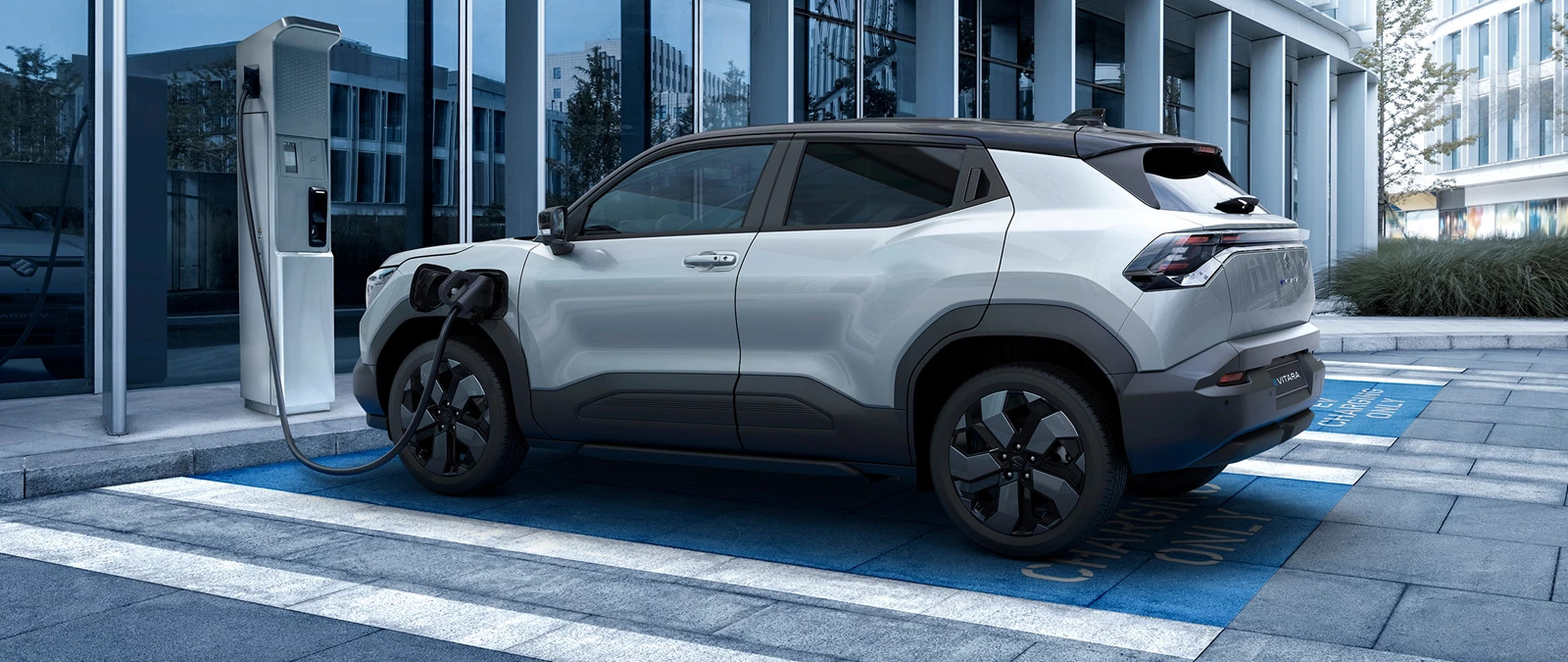 A silver Suzuki e Vitara with a black roof plugged into a charging station.